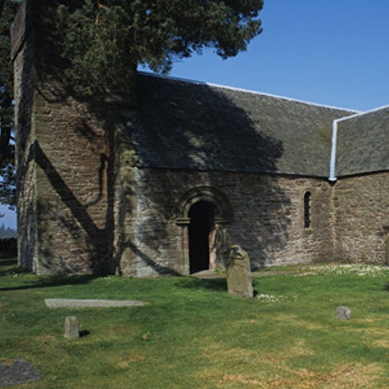 Medieval church, with grey old stone in a churchyard with some stone gravestones and a large green tree at lest of picture on a sunny day with blue skies.