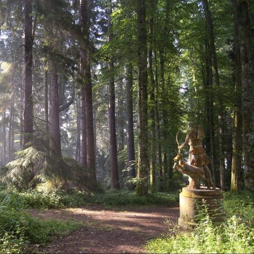 Forest path, with sunshine filtering through trees and a sculpture of a woman with a bow and arrow and a deer.