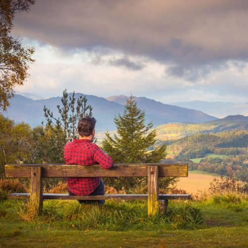 Man sitting on a wooden bench wearing a red plaid shirt looking out at a view of hills and beautiful scenery.
