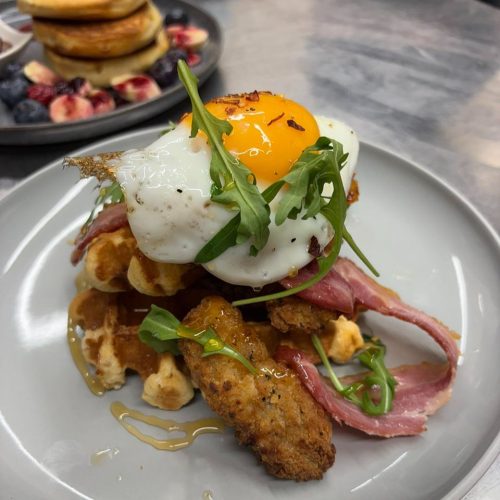 White plate with bacon, waffles and an egg garnished with rocket. Pancakes with blueberries and raspberries on a plate in background