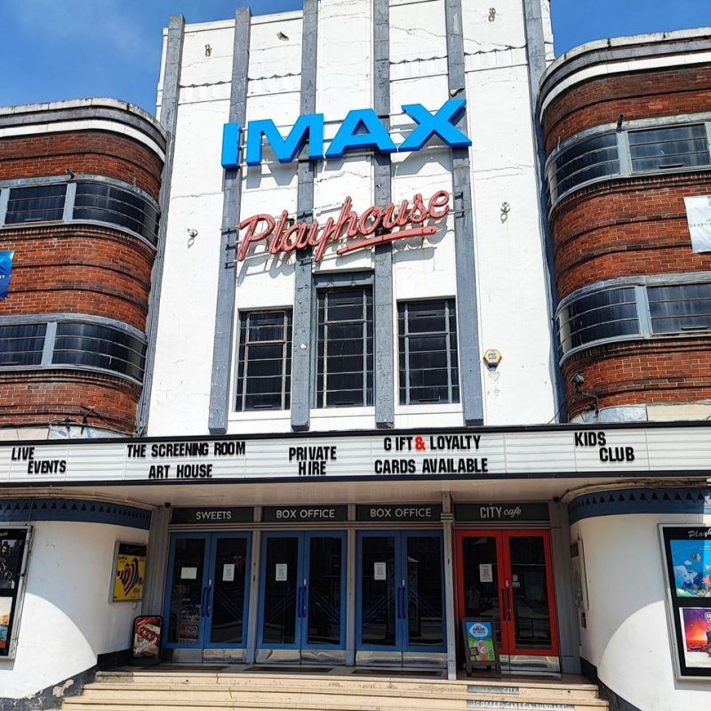 Large Art Deco building with painted white front  with blue IMAX sign, red Playhouse sign underneath and two curved red brick with windows at either side.