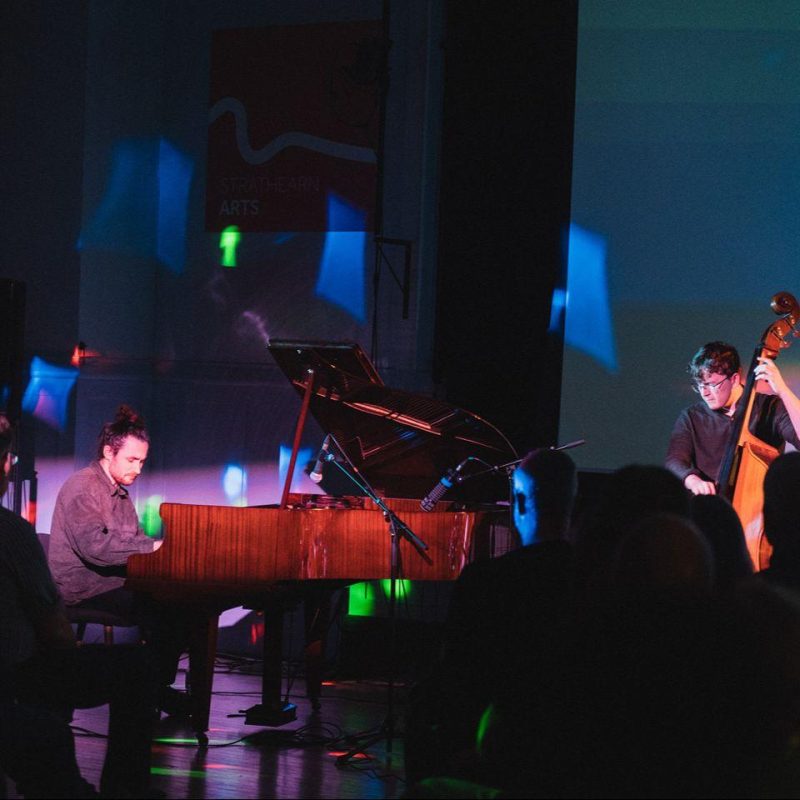 Man playing a piano on stage accompanied by a man with a chello.