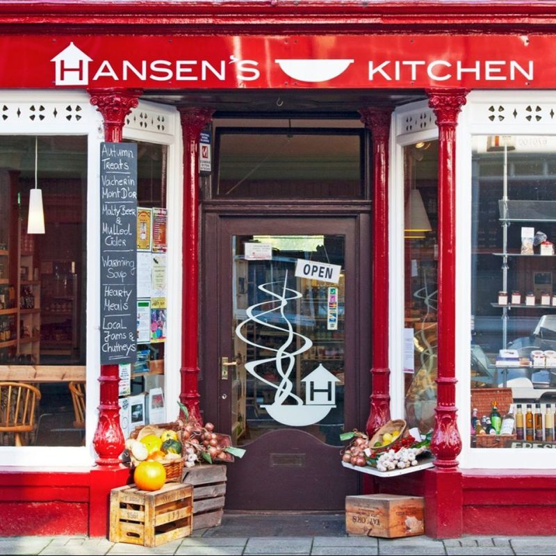 Red shop front displaying fresh veg and fruit at doorway, white trim on large windows , bar seats inside, signage Hansen's Kitchen.