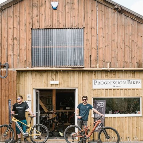 Two men with mountain bikes standing in front of wooden building with signage for Progression Bikes Dunkeld.