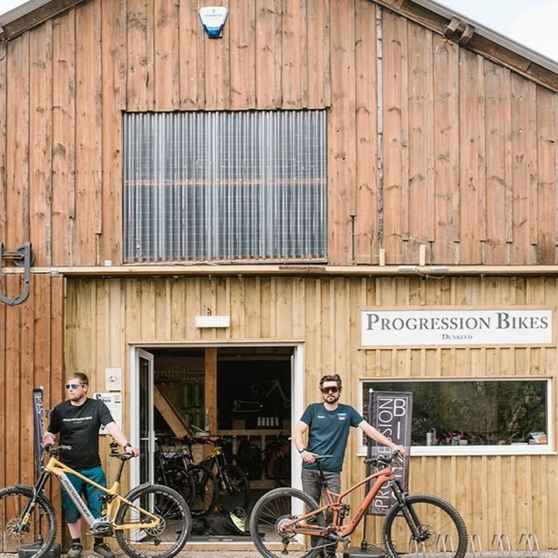 Two men with mountain bikes standing in front of wooden building with signage for Progression Bikes Dunkeld.