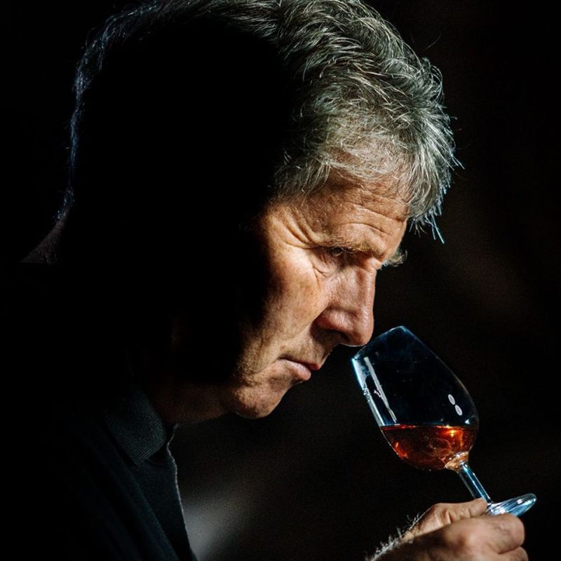 Grey haired man, smelling a glass with an amber liquid in it.