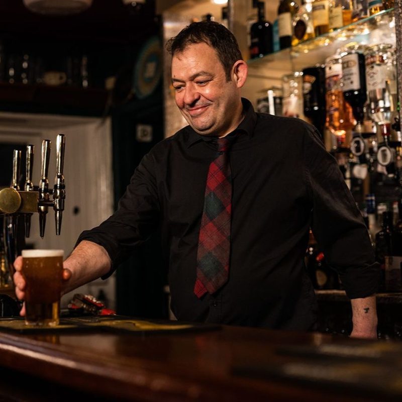 Smiling man in a black shirt behind a bar serves a lager