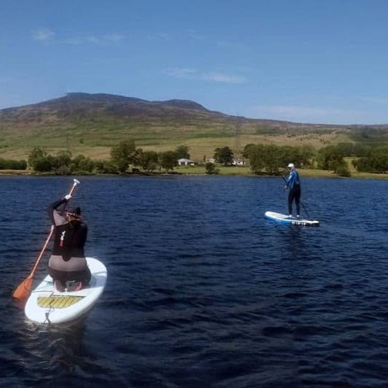 Two people on paddleboards, one kneeling, the other standing , on a loch with lush green hills and trees in the distance on a sunny day with blue skies.