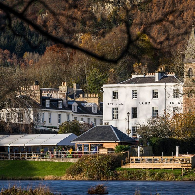 Painted white old building with The Atholl Arms Hotel signage, wooden gazebos with people sitting, in front of hotel, beside river, surrounded by trees in shades of green and brown