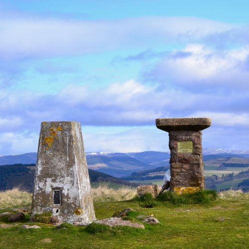 Two stone markers, green grass with green hills with cloudy blue skies.