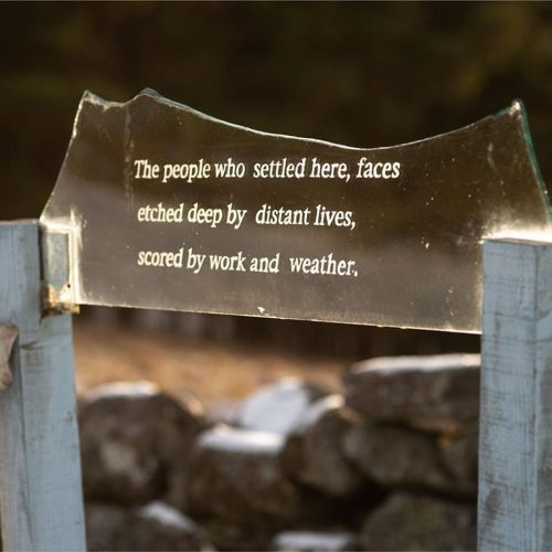Glass etched with "the people who settled here, faces etched deep by distant lives, scored by work and weather" on wooden posts with a stone wall behind.