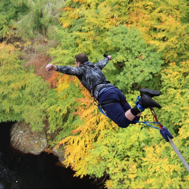 Man with bungee cord attached to ankles. over a  body of water and trees at the side.