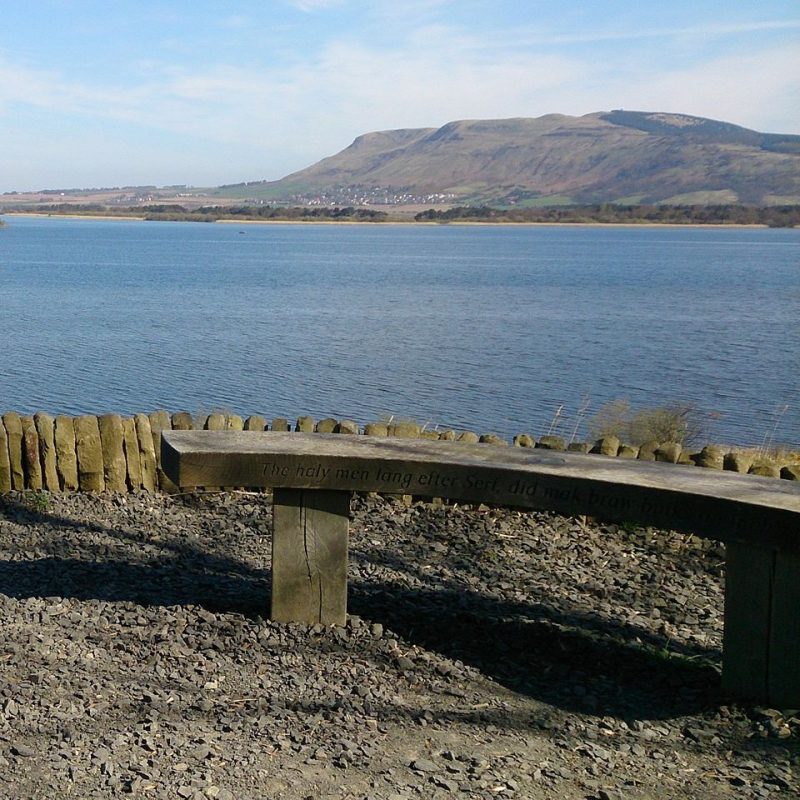 Small rounded wooden bench overlooking the body of water that is Loch Leven with hills in the distance on a sunny day with blue skies.