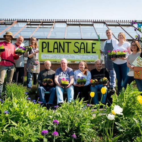 Smiling group of people holding plants in a garden in front of a greenhouse with a sign saying plant sale