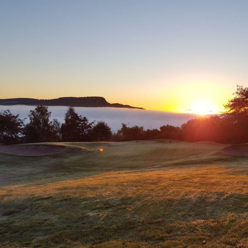 View of a green golf course with level of white clouds and hills and the sun rising in distance.