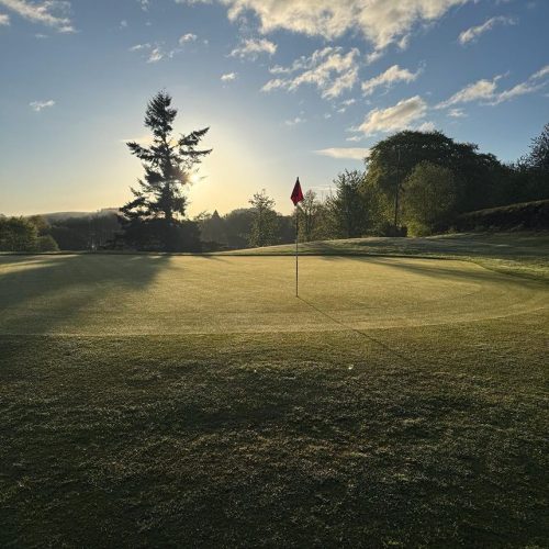 Golf hole with red flag on at dusk with green trees surrounded the course