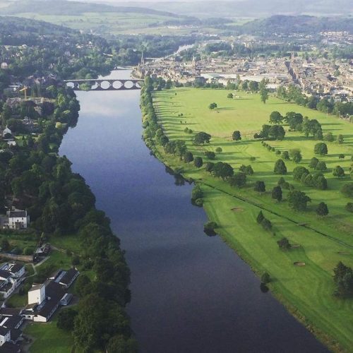 Ariel view of City of Perth, Scotland with River Tay in middle, right hand side there is a green golf course with trees and town buildings in distance.