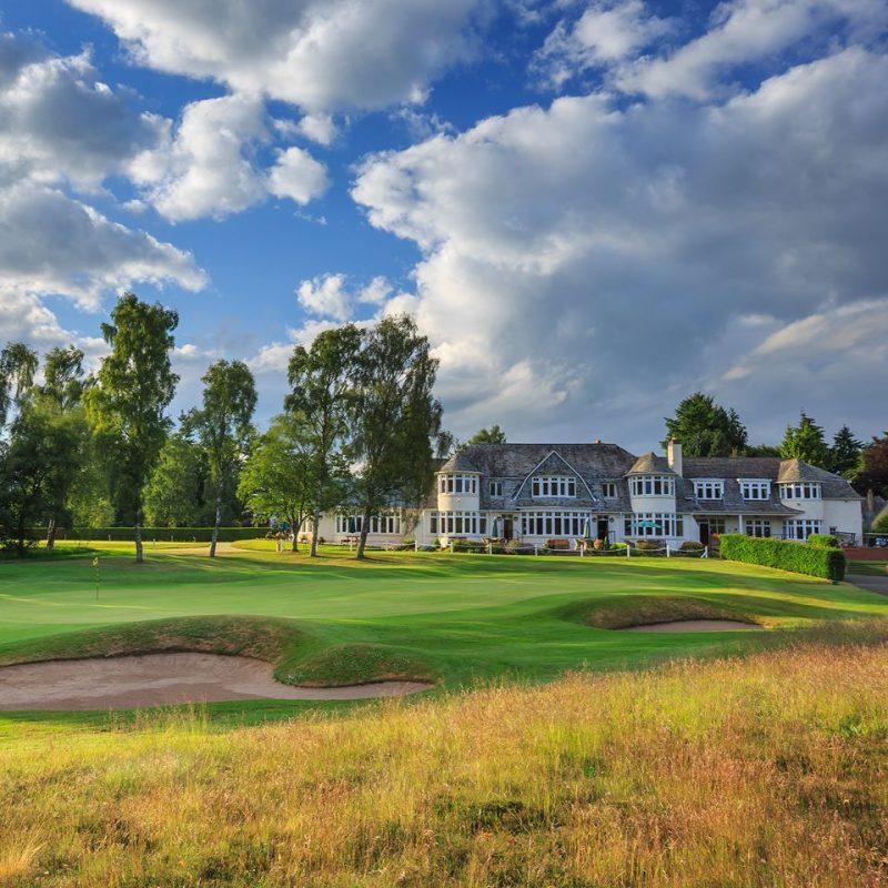 Yellow grass leading to a sand golf bunker, green fairway with an old white large building in the distance, beside green trees under a blue cloudy sky