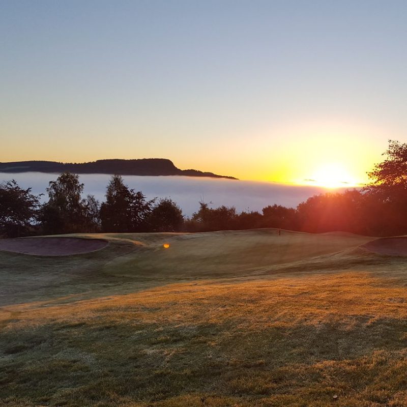 View of a green golf course with level of white clouds and hills and the sun rising in distance.
