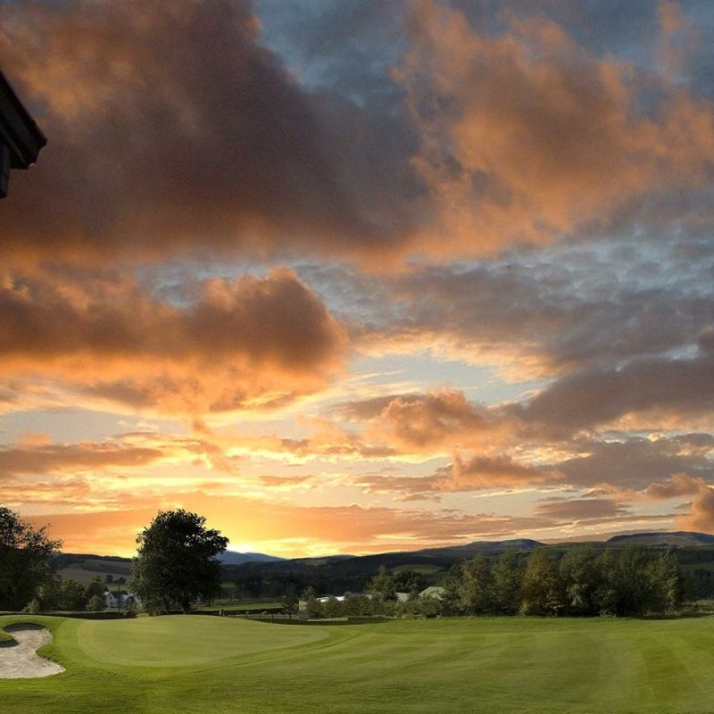 Green fairway with sand bunker to left, green trees in distance under a cloudy, sunset sky.