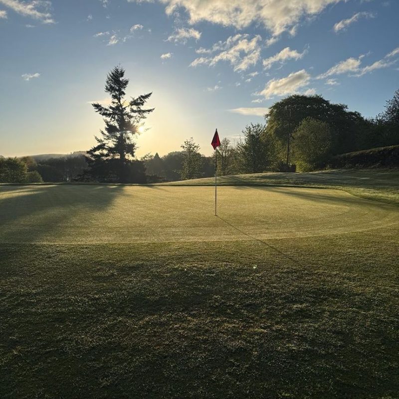 Golf hole with red flag on at dusk with green trees surrounded the course
