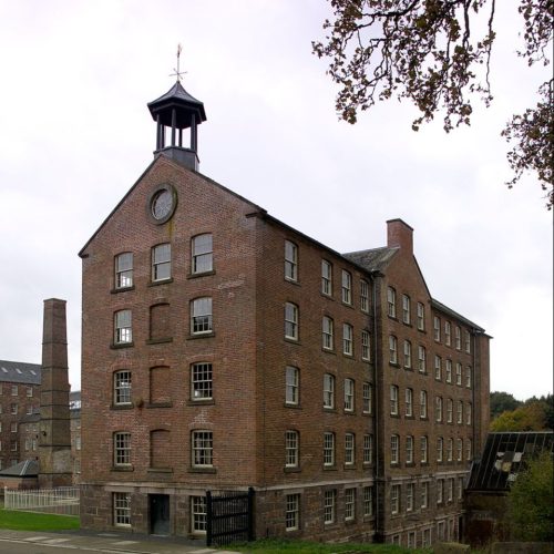 Large Victorian, red brick building with lots of windows, large industrial chimney in background, and other buildings in background.