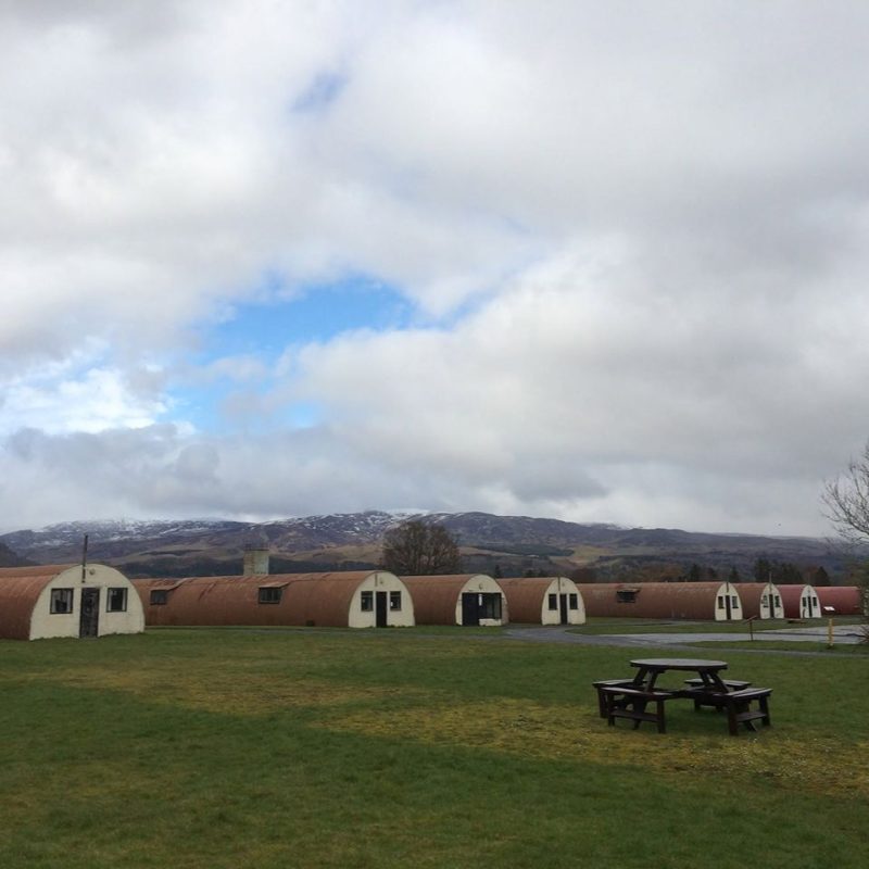 Picnic bench on grass in foreground with eight semi-cylindrical buildings with corrugated iron roofs and white-painted gables in distance, with hills behind and a cloudy sky.