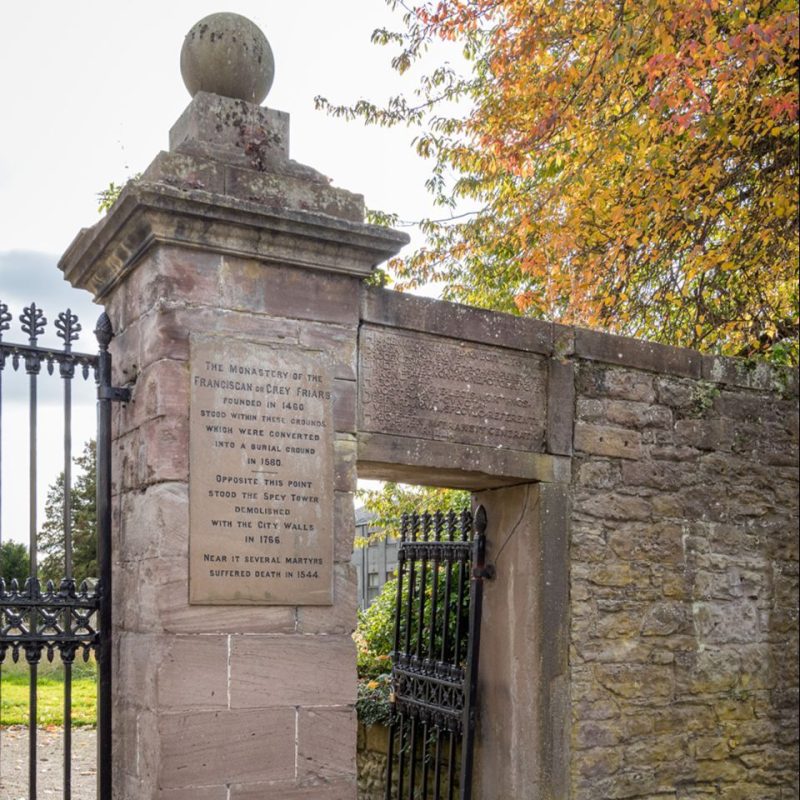 Old stone wall and gate entrance with black cast iron gate, plaque carved Monastery of Greyfriars founded in 1460 which was replaced by Greyfriars Burial Ground in 1580. with an autumnal tree, leaves on right hand side .