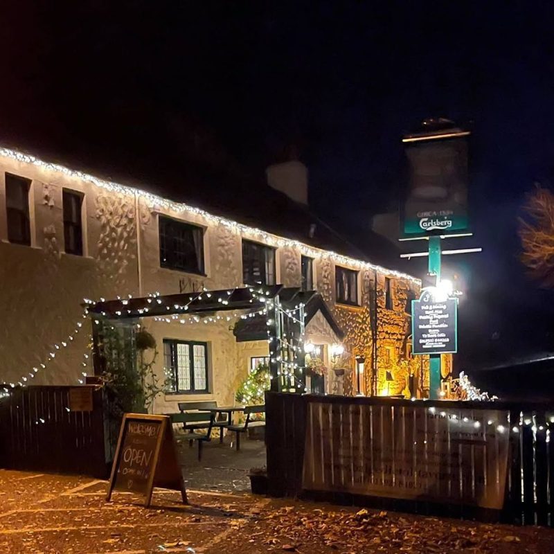 Old white painted building under a night sky with a wooden fence and entrance , hung with lights and signage for Balgedie Toll Tavern