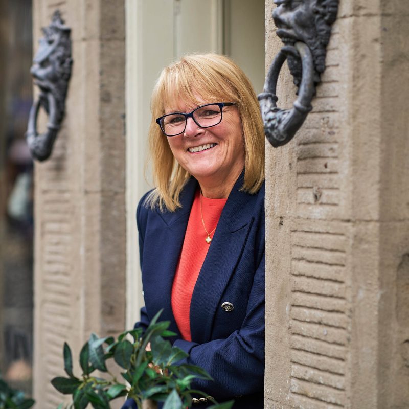 Smiling blonde haired lady wearing a navy jacket in a doorway with two iron ornate door knockers.