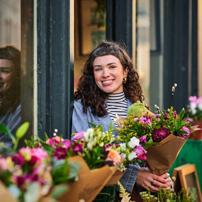 Smiling woman with dark curly hair , standing in a doorway holding a bunch of colourful flowers. Bunches of flowers in foreground.
