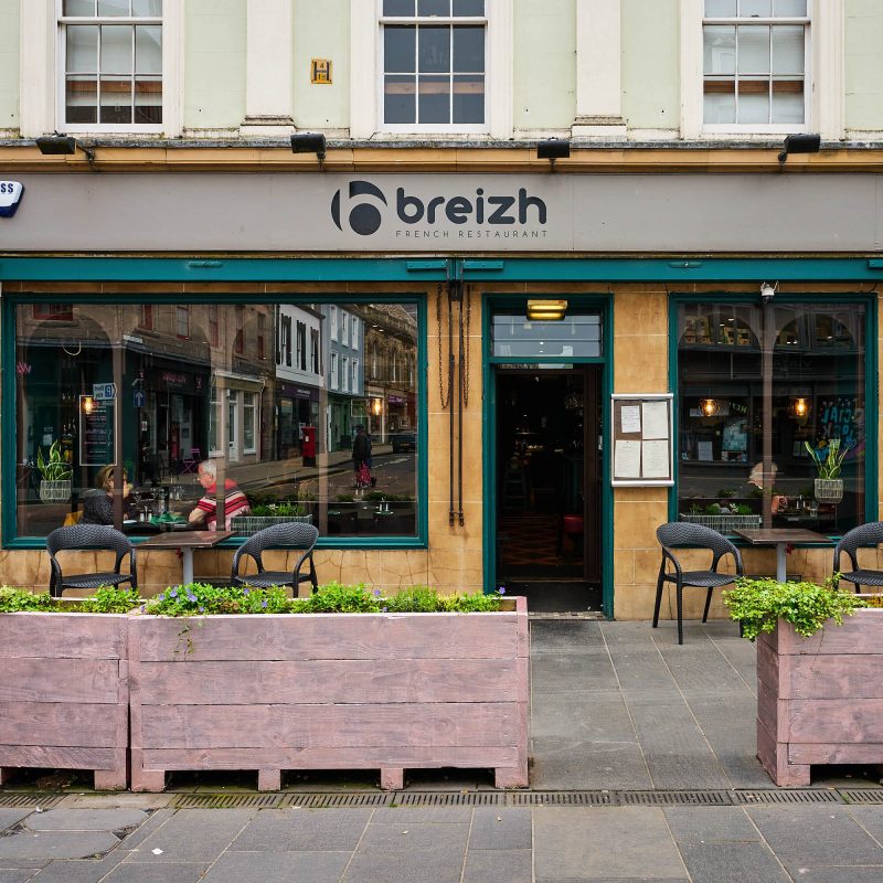 Shows a brick building with teal accents around the windows. Above is a grey sign which says 'Breizh French Restaurant' in black text. In front of the restaurant is wooden boxes filled with flowers.