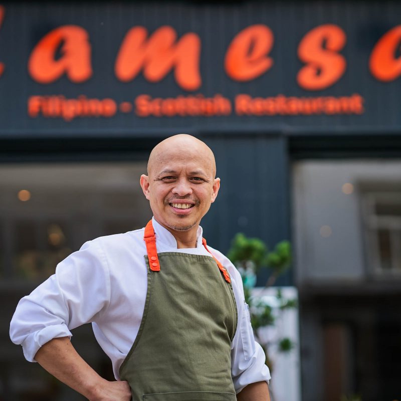 A smiling man wearing a white chefs jacket and green apron with orange straps is standing in front of a grey building with an orange sign which says 'Lamesa'
