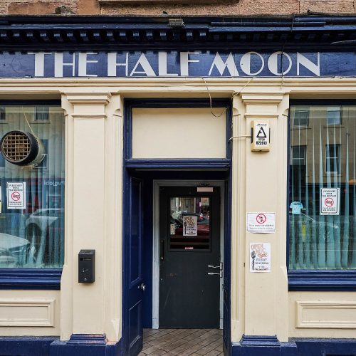 A beige building with dark blue accents around the windows and the door, There is a sign which say is grey text 'The Half Moon'