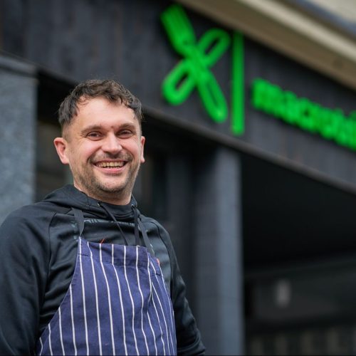 Smiling man with blue and white apron stands below a shop sign for Macrotrition in Perth, Scotland