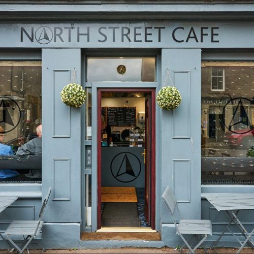 Grey painted shop front, North street cafe painted in black above door, tables and chairs either side of doorway.