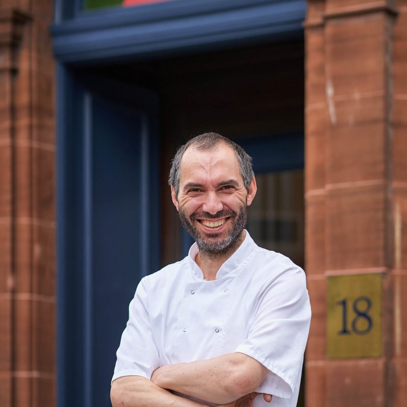 A smiling man wearing a white chefs jacket standing outside a brick building with a blue door