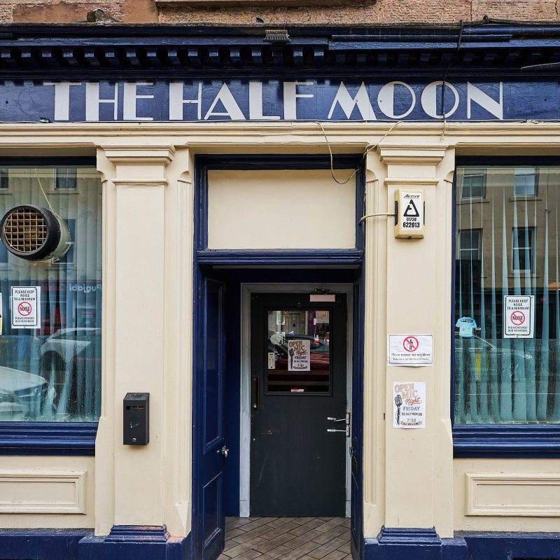 A beige building with dark blue accents around the windows and the door, There is a sign which say is grey text 'The Half Moon'