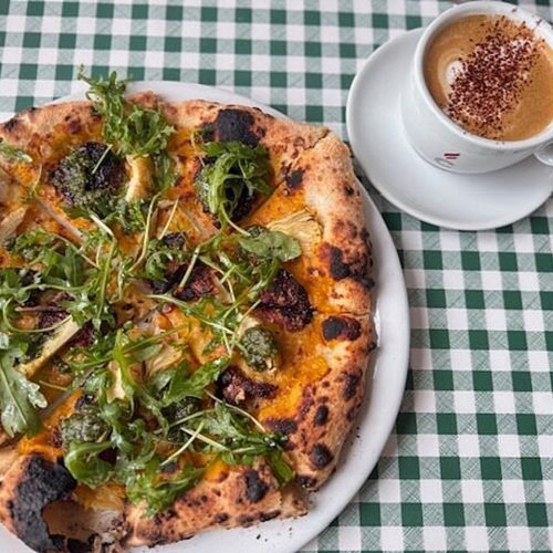 Pizza with meats and cheese, topped with rocket and a white cup and saucer filled with coffee, sitting on a green and white check tablecloth.