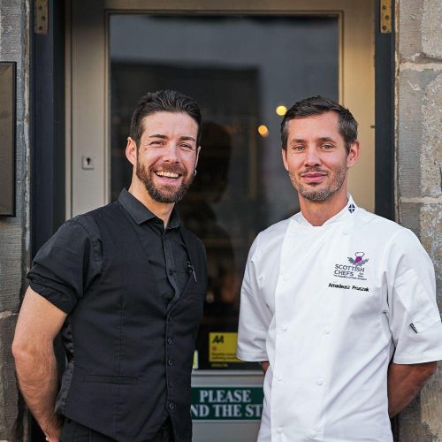 Two smiling men, one in a black top and one in chef whites