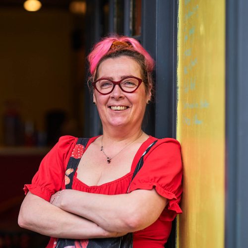 A smiling women wearing a red top is standing cross armed leaning against a yellow wall