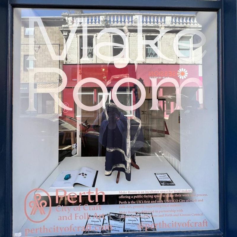 Window with Make Room and Perth City of Craft and Folk arts decals, blue and white scarf displayed behind glass.