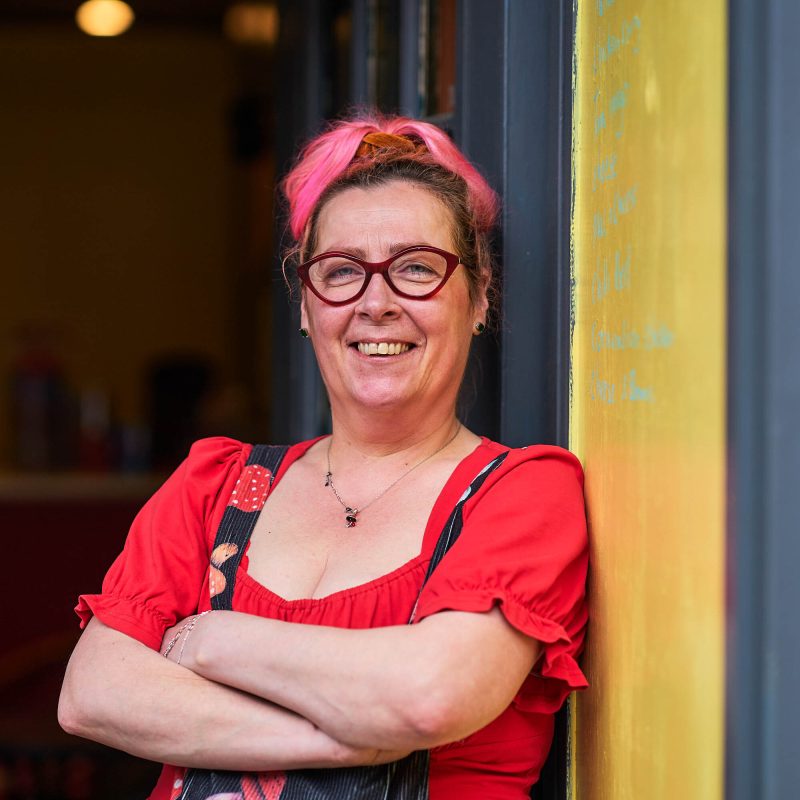 A smiling women wearing a red top is standing cross armed leaning against a yellow wall