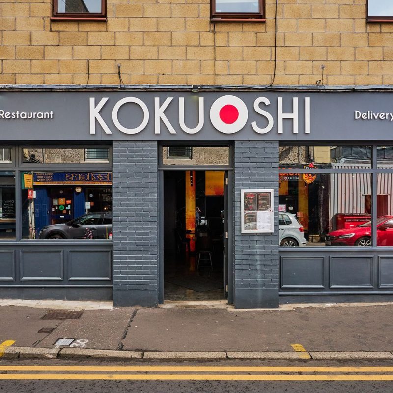 Grey painted shop front with Koku Shi in white letters, white circle with red circle inside between Koku and Shi.