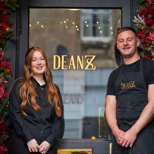 One man and a woman with long red hair, wearing black, man wears an apron with Deanzs logo,in front of a door with Deanzs logo in gold.