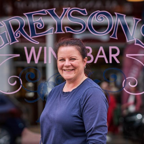 A smiling lady in a navy t-shirt standing in front of a glass window which has the text 'Greysons Wine Bar' on it in pink letters.