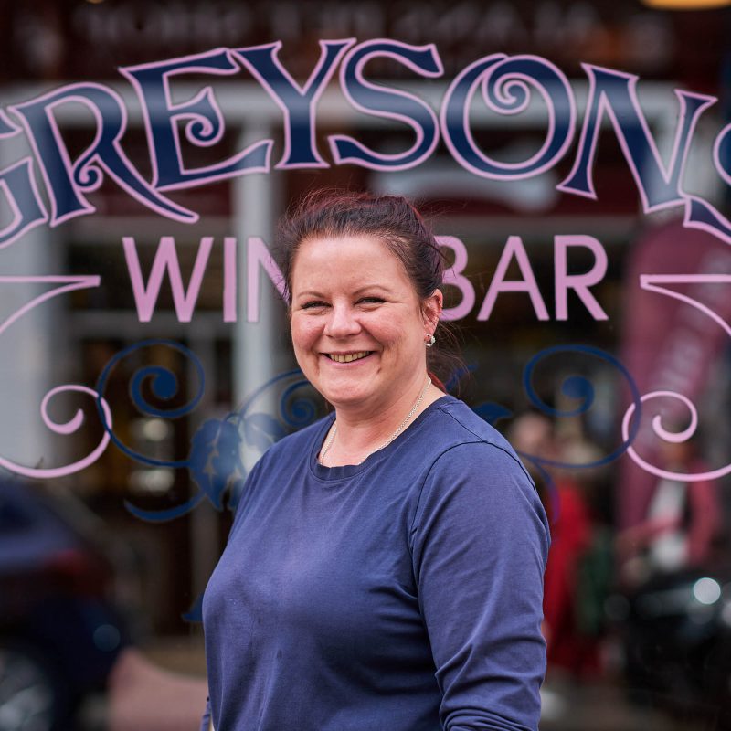 A smiling lady in a navy t-shirt standing in front of a glass window which has the text 'Greysons Wine Bar' on it in pink letters.