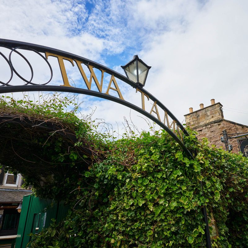 A black sign with gold letters which read 'Twa Tams' going over a pathway which is surrounded by bushes.