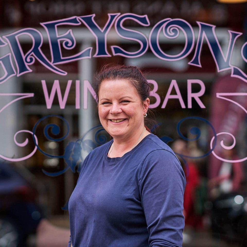 A smiling lady in a navy t-shirt standing in front of a glass window which has the text 'Greysons Wine Bar' on it in pink letters.