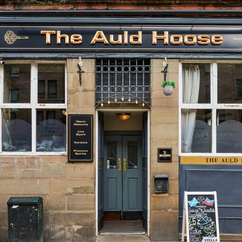 A stone building with grey-blue doors. Above the door is a black sign with gold letters which say 'The Auld Hoose.'